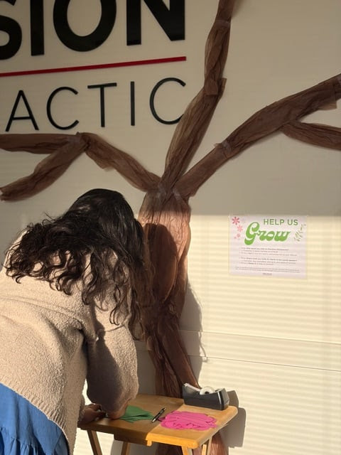 A patient leans over a table to write a referral for the referral tree, which is brown tissue paper taped onto the wall in the shape of a tree.